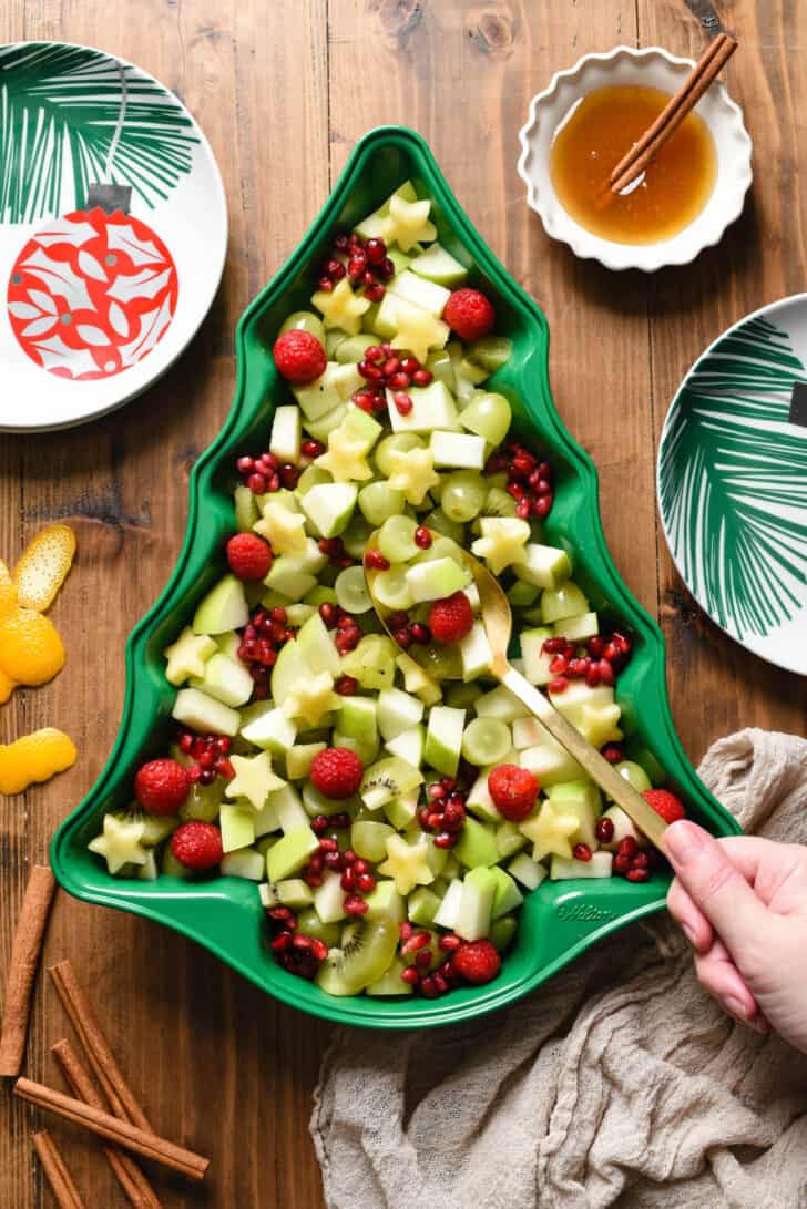 Christmas fruit salad made with grapes, raspberries, pomegranate seeds, kiwi, pineapple and green apple, arranged in a green Christmas tree shaped baking pan, with a woman's hand using a gold spoon to scoop some out.
