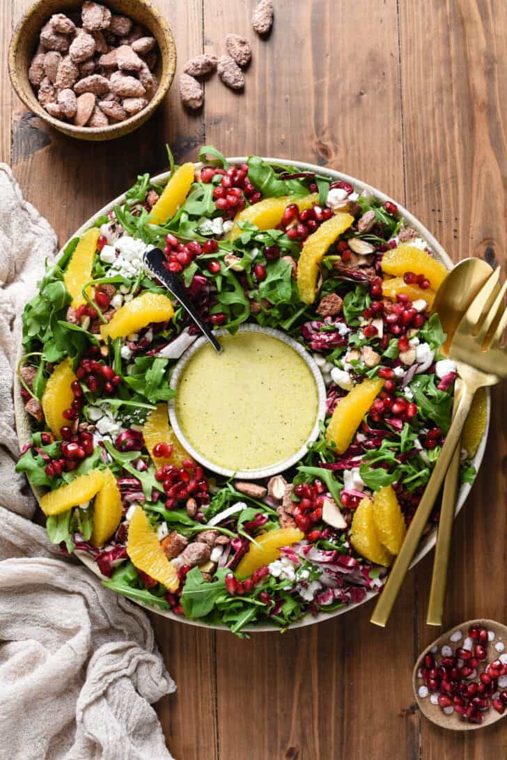 A Christmas Salad recipe made with greens, oranges, cheese, pomegranate seeds and nuts, arranged like a wreath with a bowl of dressing in the middle.