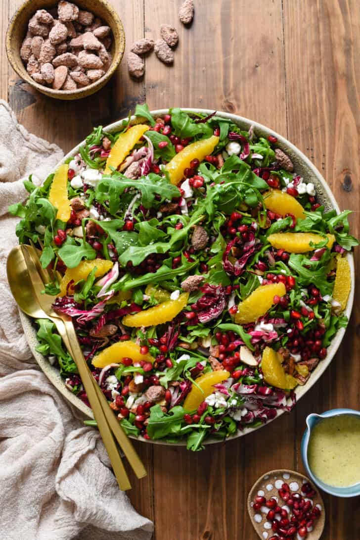 A salad for Christmas dinner with greens, oranges, cheese, pomegranate seeds and nuts, arranged on a large circular platter.