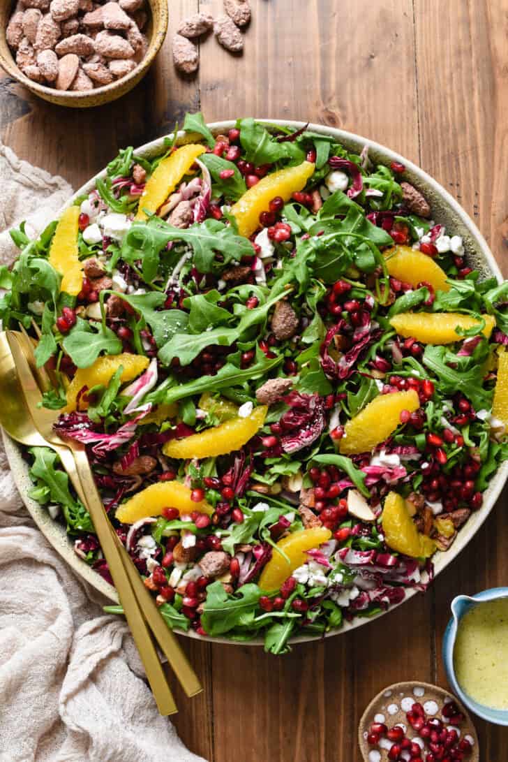 A Christmas Salad recipe made with greens, oranges, cheese, pomegranate seeds and nuts, arranged in a large bowl with gold salad servers perched on side of bowl.
