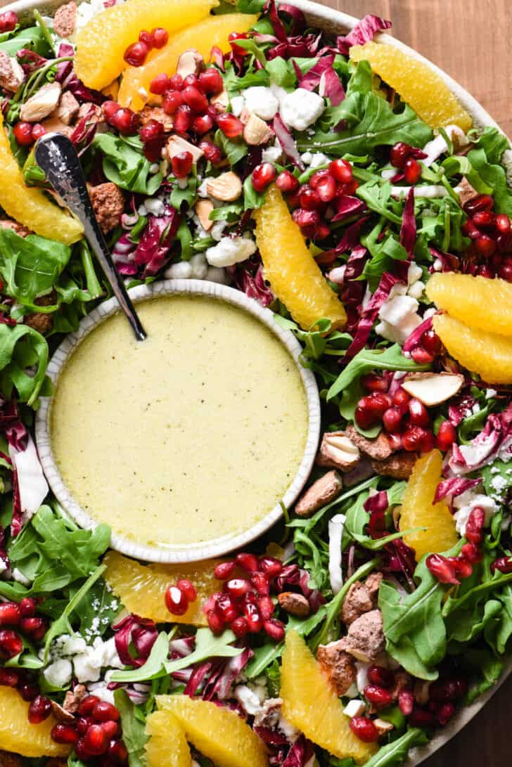 An arugula goat cheese salad made with oranges, pomegranate seeds and nuts, arranged in a large bowl with a smaller bowl of orange salad dressing.