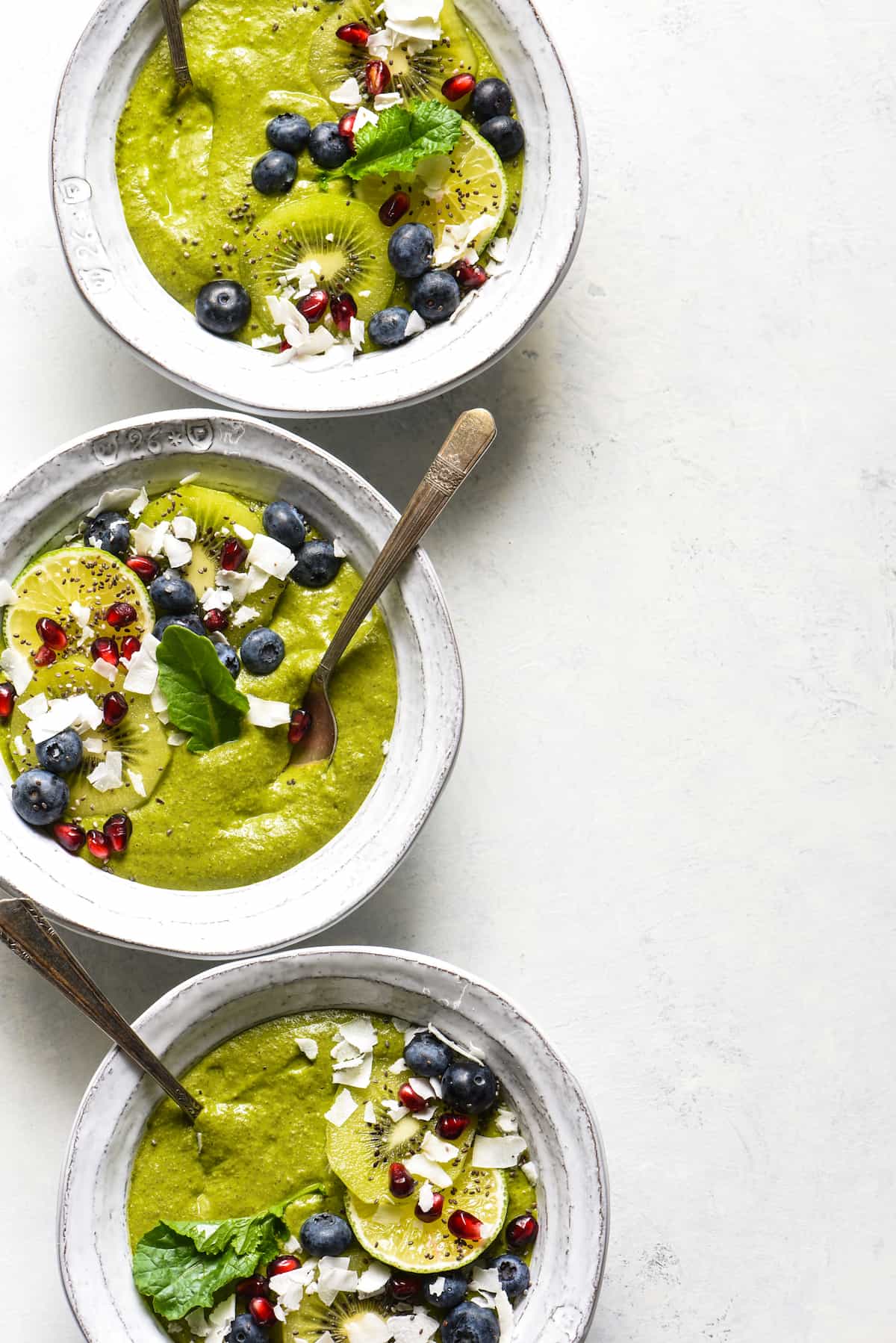 Three tropical smoothie bowls topped with blueberries, pomegranate seeds, coconut, kiwi and a green leaf.