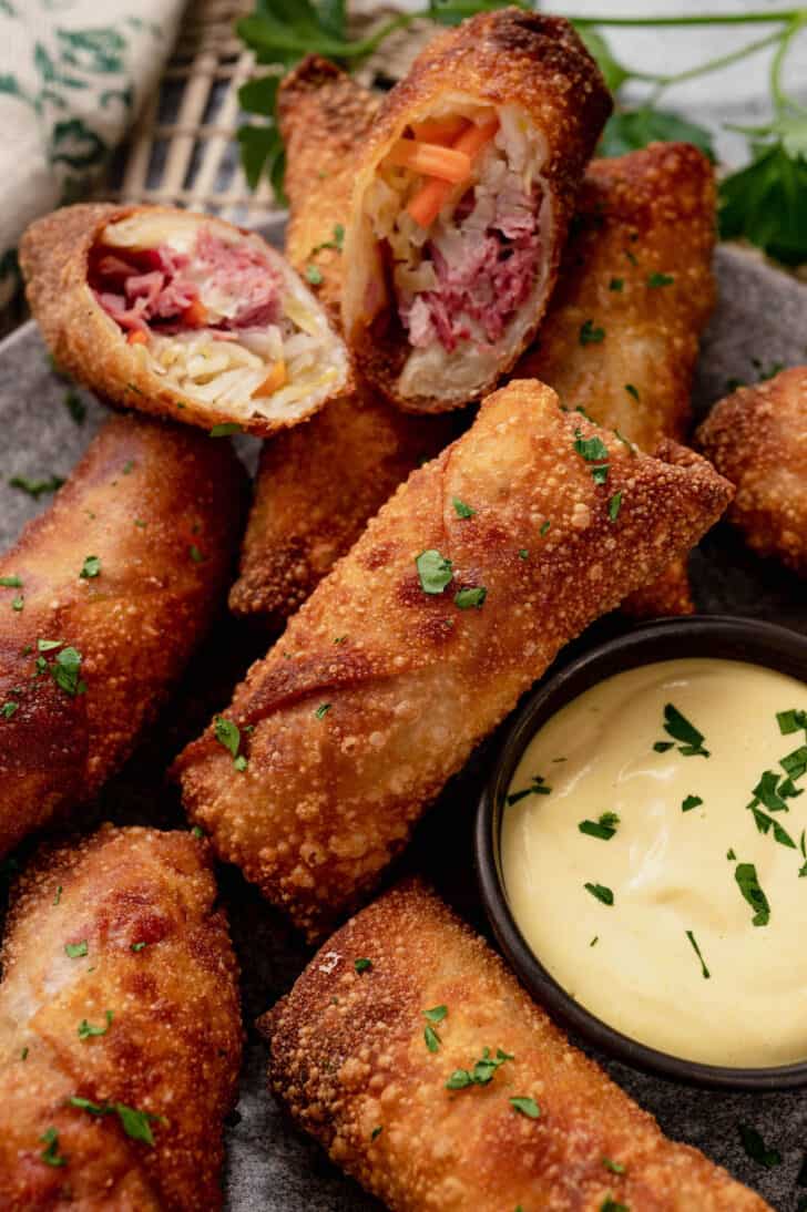 A plate of corned beef egg rolls with creamy mustard dipping sauce. One egg roll is cut in half, showing the inside filling.