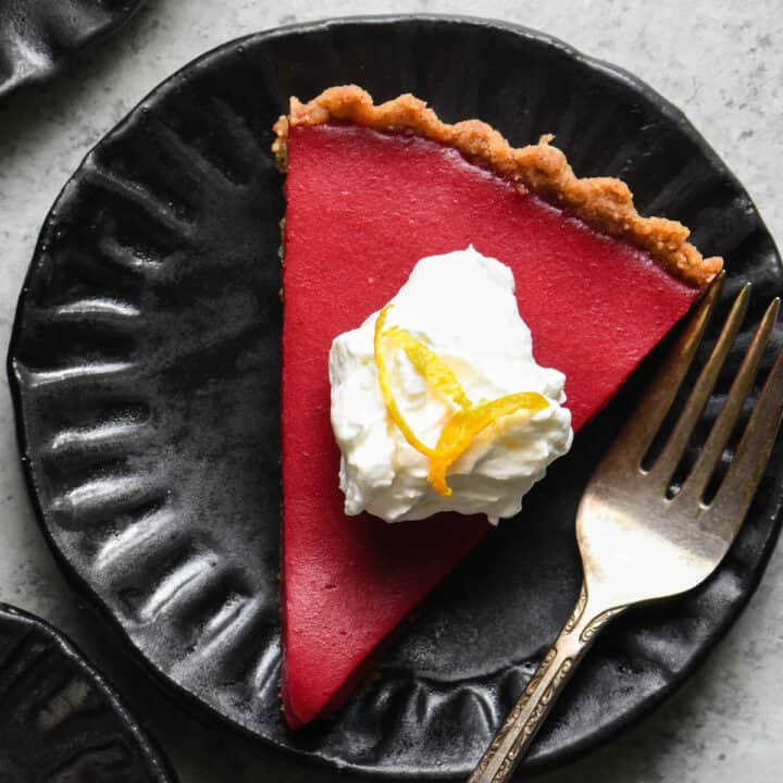 Overhead photo of a slice of cranberry curd tart on black plate, topped with whipped cream and orange zest.