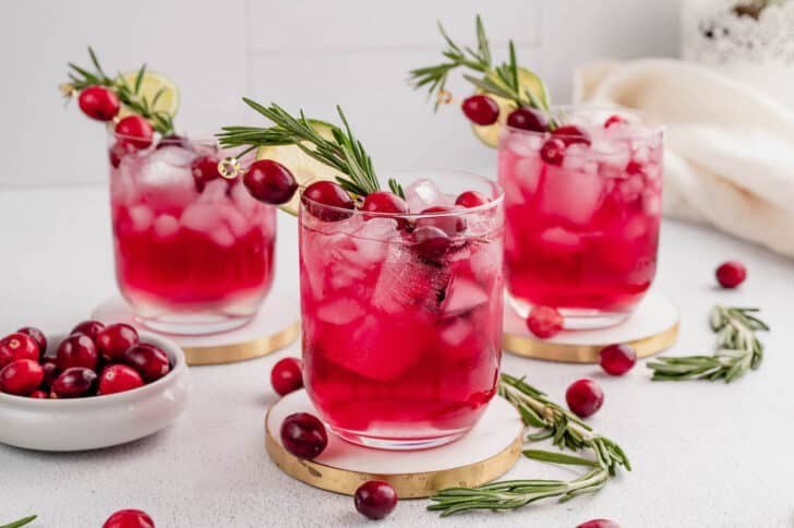 Three Christmas cranberry cocktails resting on wooden coasters on a light surface.
