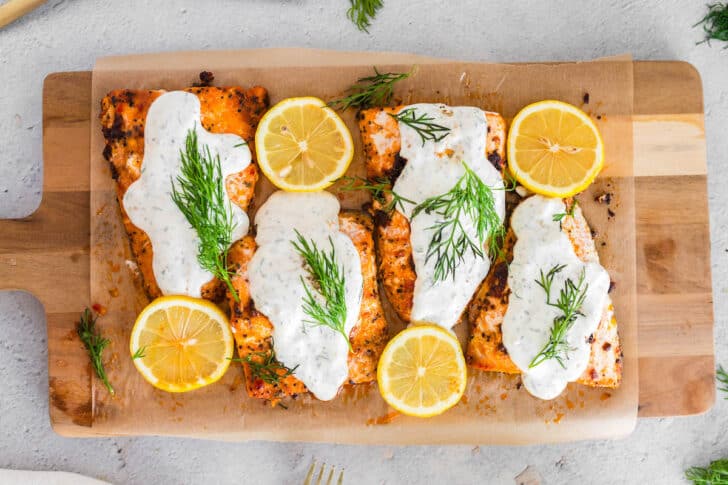 Four fish fillets on a cutting board topped with lemon dill sauce for salmon.
