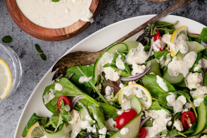 A large white platter filled with salad made from mixed greens, cucumber ribbons and grape tomatoes, tossed with feta dressing.