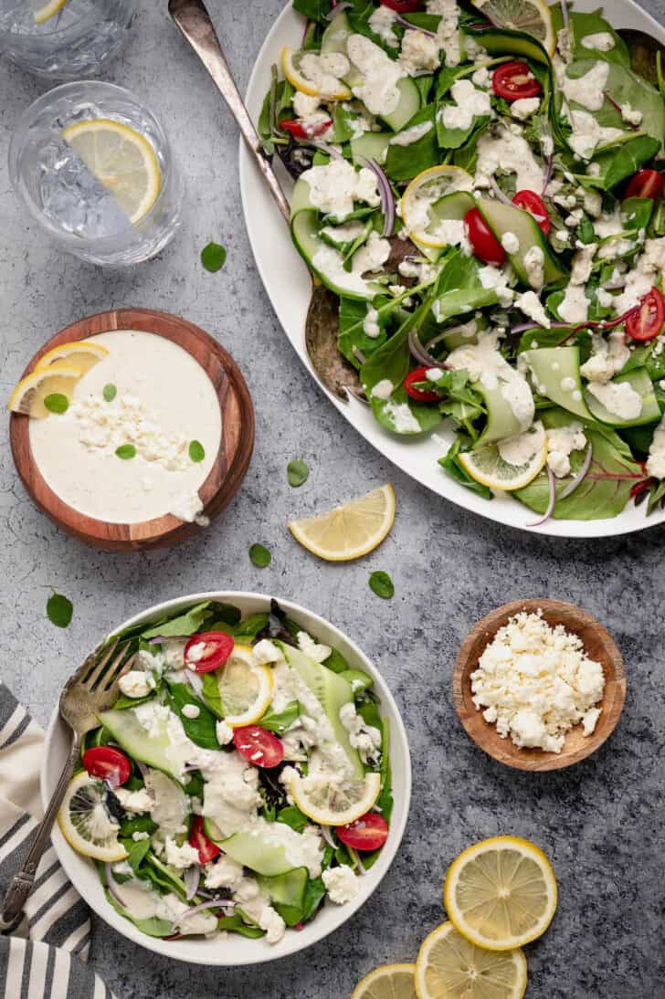 A large white platter and a smaller bowl filled with salad made from mixed greens, cucumber ribbons and grape tomatoes, tossed with feta dressing.