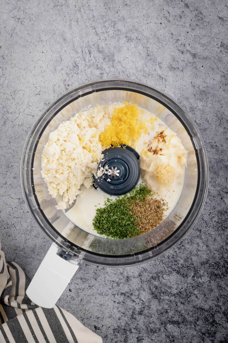 A food processor bowl filled with the ingredients for feta salad dressing, including feta cheese, lemon zest, garlic, milk, mayonnaise, herbs and spices.