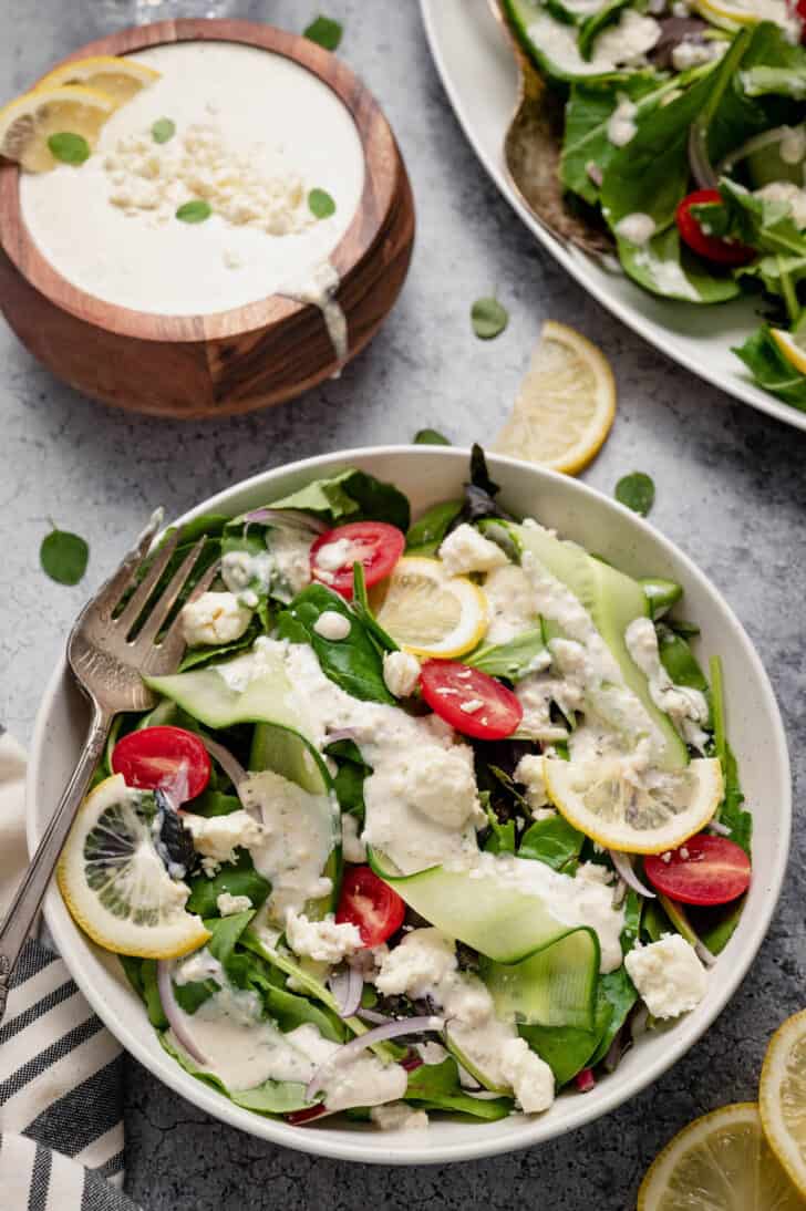 A white bowl filled with salad made from mixed greens, cucumber ribbons and grape tomatoes, tossed with creamy feta dressing, with a wooden bowl of dressing in the background.