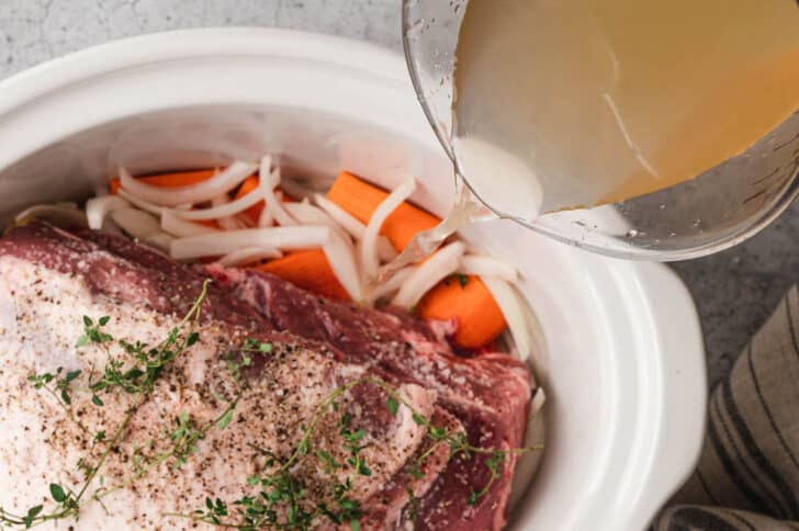 A white slow cooker insert with sliced carrots, onions and garlic with a pork roast on top inside, with broth being poured in.