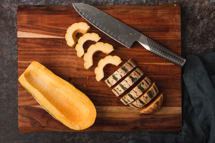 A wooden cutting board and a knife, with a fall gourd being slices into half moon slices.