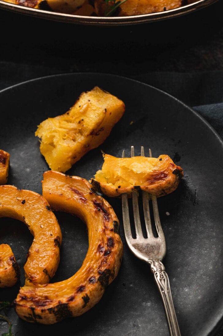 A fork stabbing a piece of roast delicata squash on a black plate.