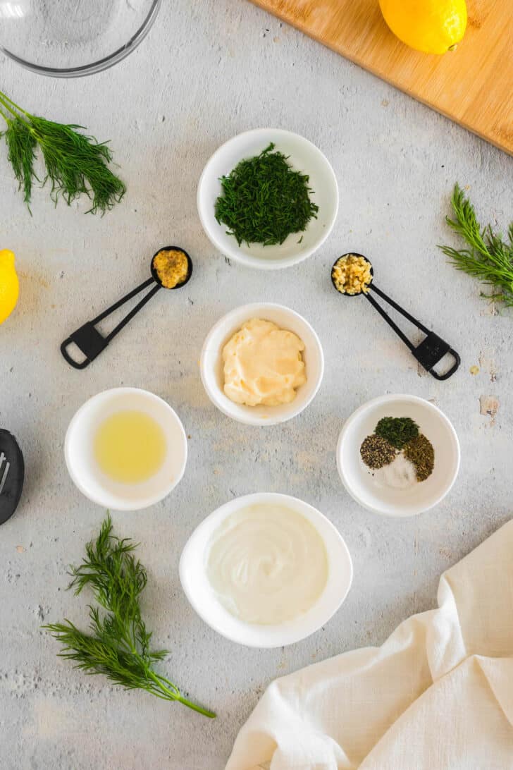 Ingredients laid out on a white surface in bowls and spoons, including herbs, garlic, mustard, yogurt and mayonnaise.