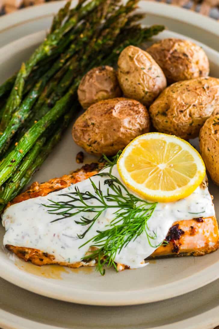 A plate of fish, asparagus and potatoes. The fish is topped with a creamy herb sauce and a lemon slice.