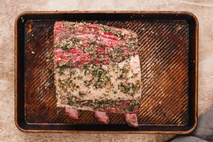 A raw standing rib roast with a rub of prime rib on it, on a rimmed baking pan.