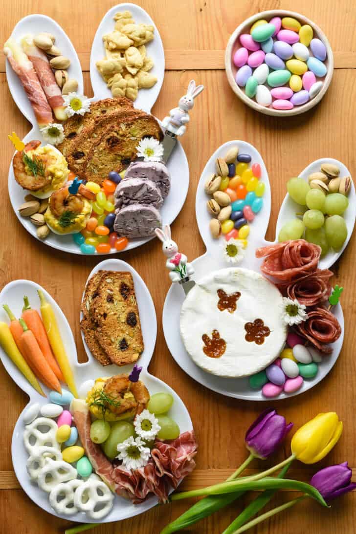 An Easter charcuterie board arranged on three white bunny head shaped plates. The plates are filled with charcuterie, cheeses, crackers, fruit, carrots, and candy.
