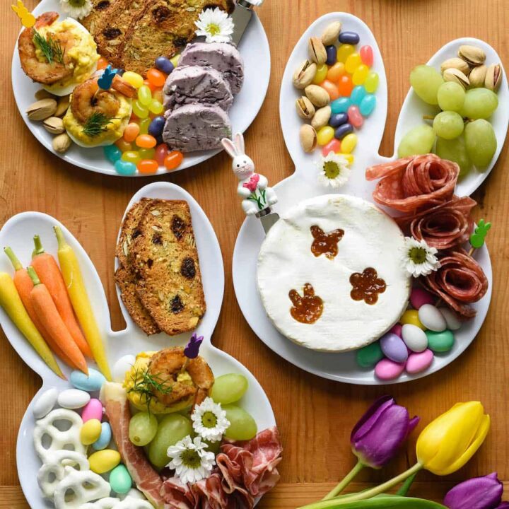 An Easter charcuterie board arranged on three white bunny head shaped plates. The plates are filled with charcuterie, cheeses, crackers, fruit, carrots, and candy.