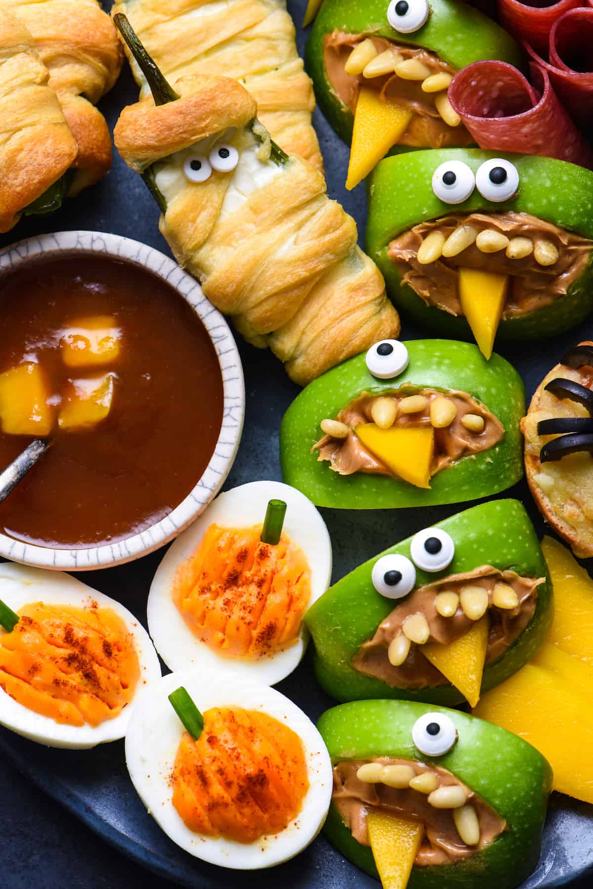 Closeup of Halloween themed food, like deviled eggs that look like pumpkins, jalapeno poppers wrapped like mummies and green apples cut out like monsters.