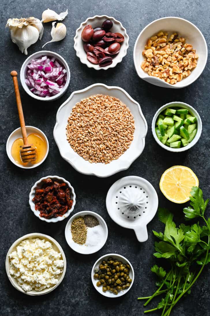 Ingredients laid out in white bowls on a dark surface, including red onion, olives, walnuts, cucumber, lemon, spices, capers, feta cheese, sun-dried tomatoes, parsley, honey, garlic and a bowl of grains.
