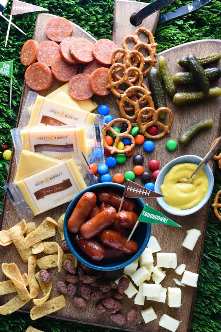 A tailgate charcuterie board made on a wooden cutting board, with cheese, sliced pepperoni, pickles, pretzels, candy, mustard, corn chips and cocktail weenies.