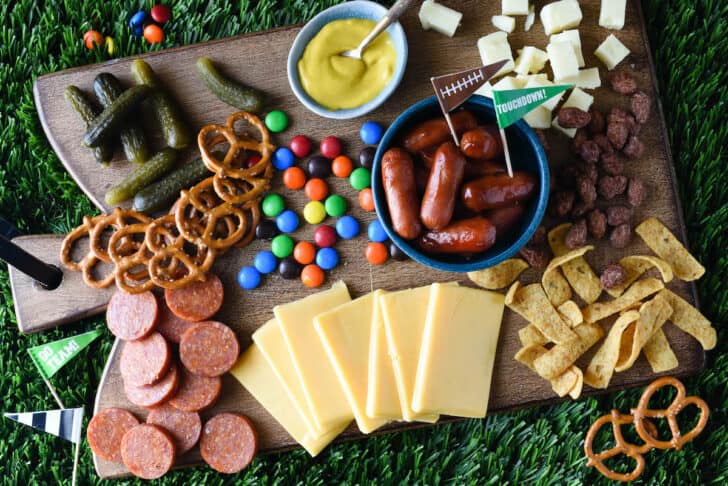 A football themed charcuterie board made on a wooden cutting board, with cheese, sliced pepperoni, pickles, pretzels, candy, mustard, corn chips and cocktail weenies.