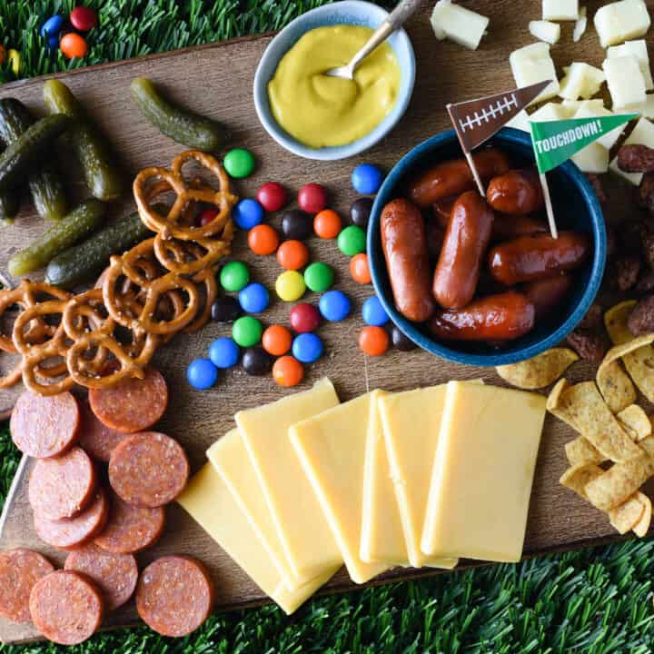 A football charcuterie board made on a wooden cutting board, with cheese, sliced pepperoni, pickles, pretzels, candy, mustard, corn chips and cocktail weenies.