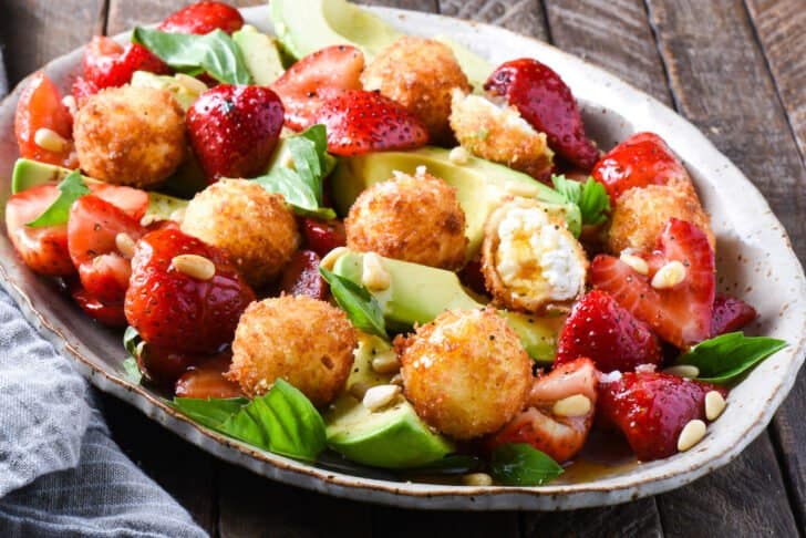Fried goat cheese salad with strawberries, avocado, pine nuts and basil, on a speckled oval plate.