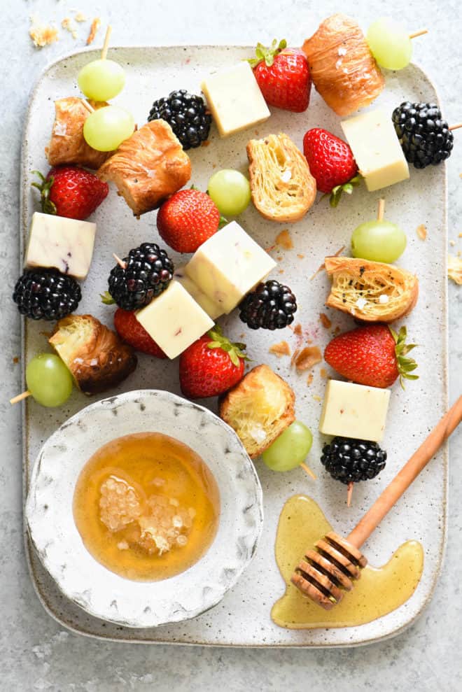 Skewers of fresh berries, white cheese and croissant pieces, with a small bowl of honey.