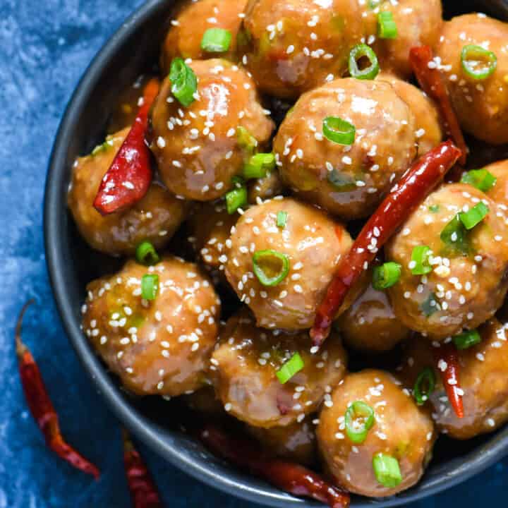 Round bowl of General Tso's meatballs topped with green onions, sesame seeds and dried chiles on a blue background.