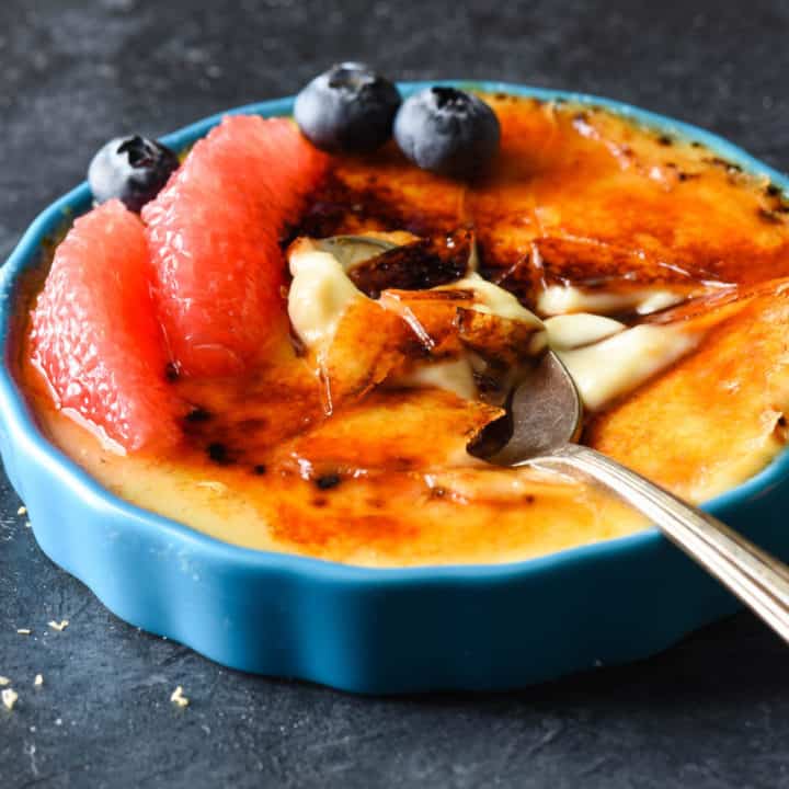 Grapefruit creme brulee in ramekins, garnished with grapefruit segments and blueberries, on a dark surface, with a spoons digging into them.