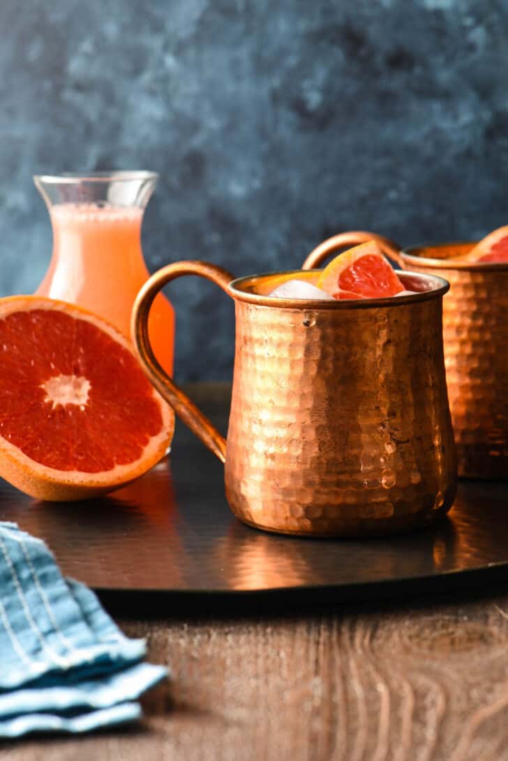 Two grapefruit mules in hammered copper mugs on a black tray with half a grapefruit and a bottle of grapefruit juice.