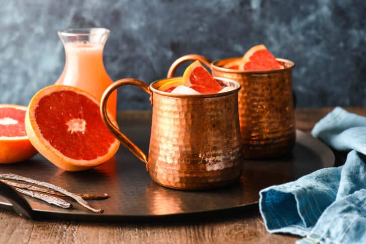 Two grapefruit vanilla mules in hammered copper mugs on a black tray with half a grapefruit and a bottle of grapefruit juice.