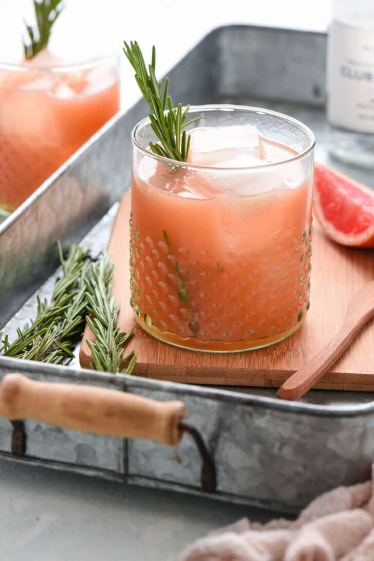 Grapefruit vodka cocktail with a rosemary sprig garnish in a textured glass on an aluminum tray.