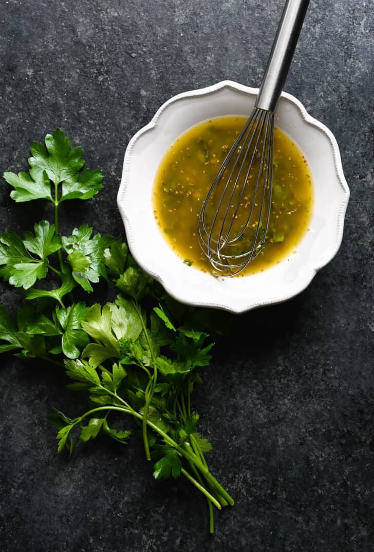 A white bowl filled with an olive-oil based dressing with a whisk in it, with a bunch of parsley alongside.