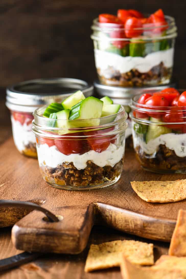 Pint sized mason jars filled with a layered Mediterranean dip, including ground lamb, yogurt, cucumbers and tomatoes.