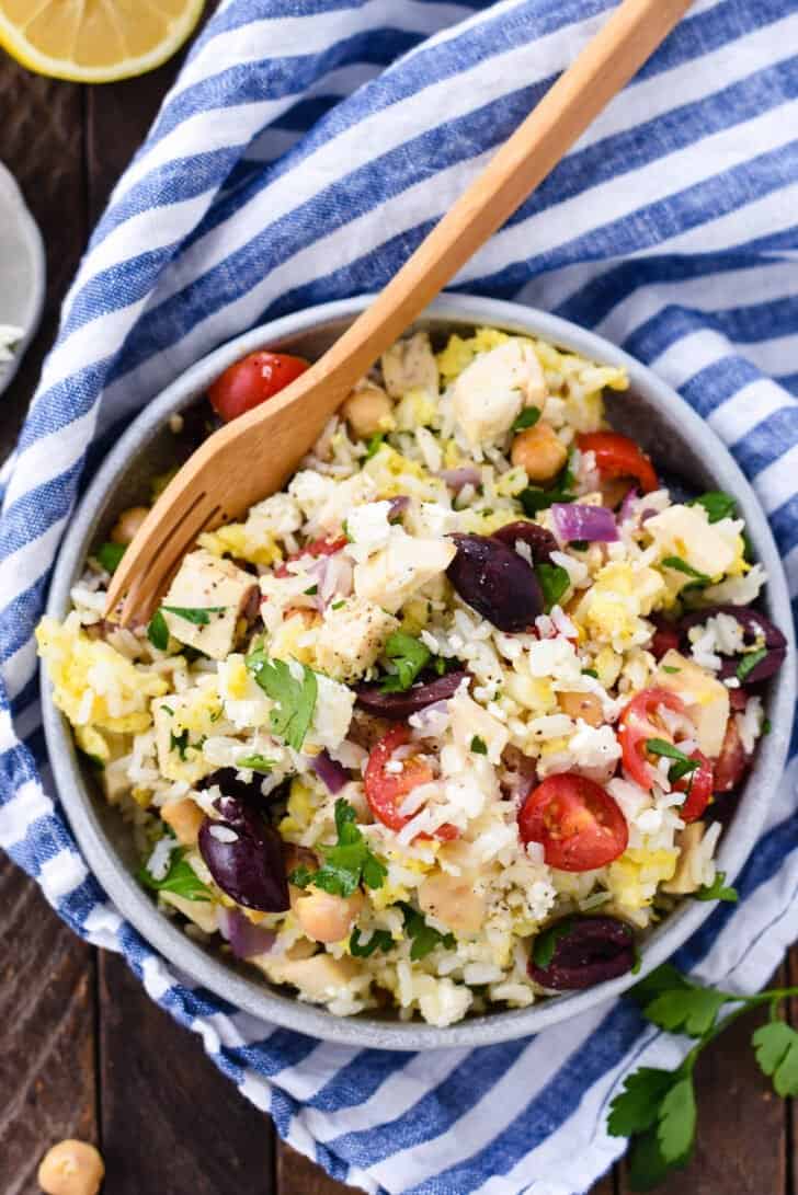 A gray bowl sits on top of a blue and white striped napkin, filled with a Mediterranean rice recipe made with chicken, tomatoes, olives, herbs and feta cheese.