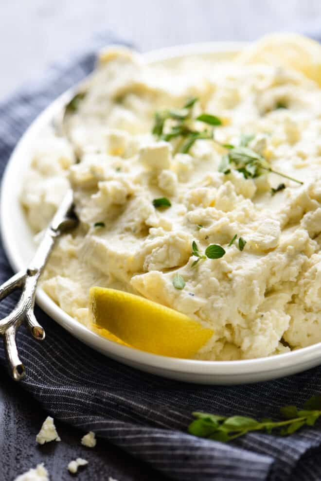 Mashed potatoes with yogurt on white platter, garnished with herbs and lemon.