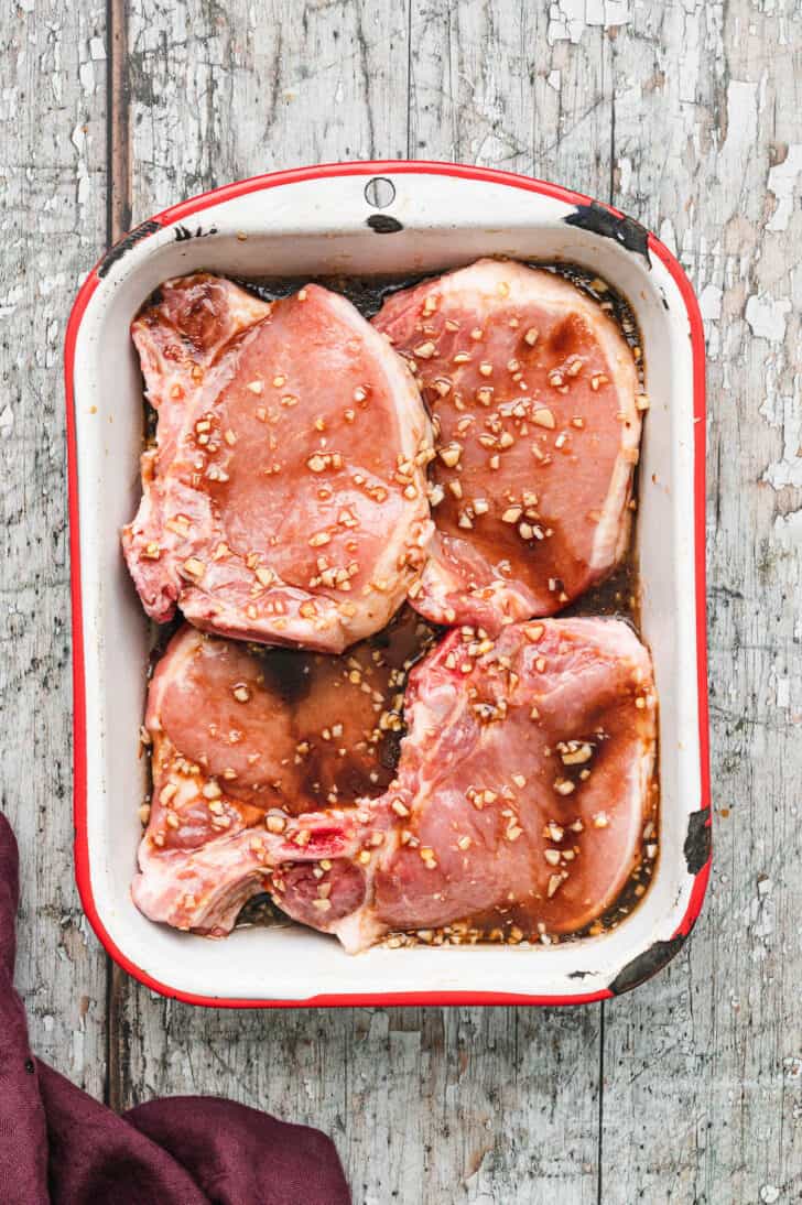 Bone in red meat covered in marinade in a rustic red and white baking dish.
