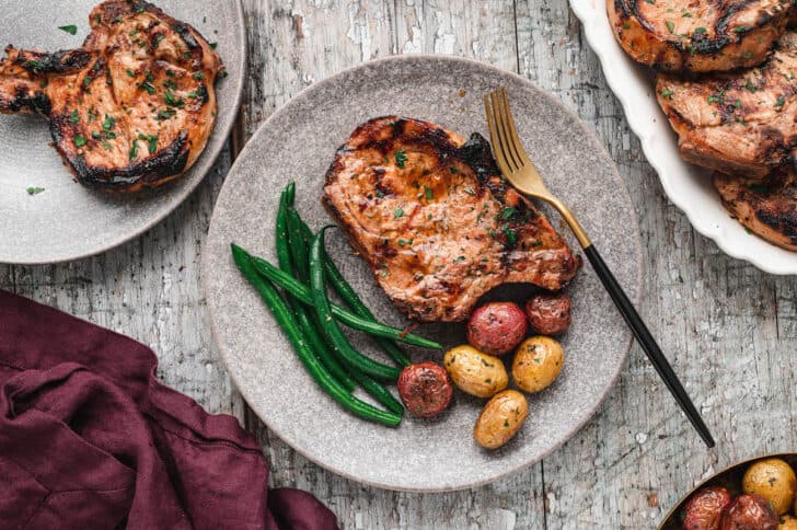 A grilled pork chop on a textured gray plate alongside roasted baby potatoes and green beans.