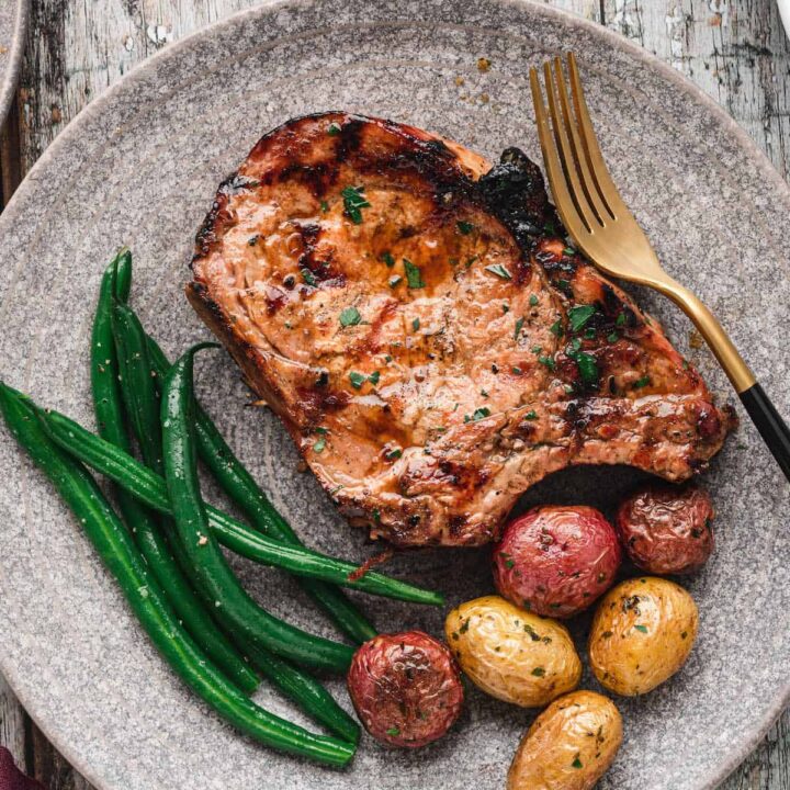 A grilled pork chop on a textured gray plate alongside roasted baby potatoes and green beans.