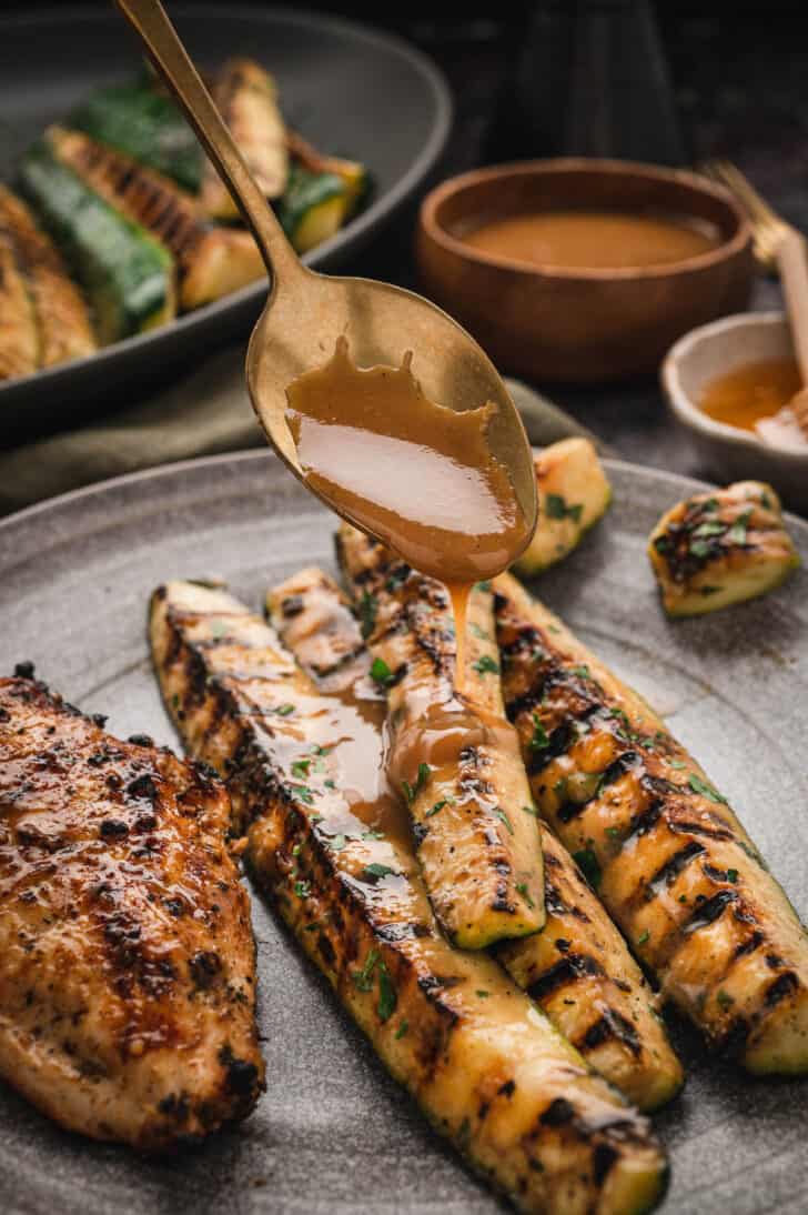 A gold spoon drizzling a light brown dressing over grilled zucchini spears.