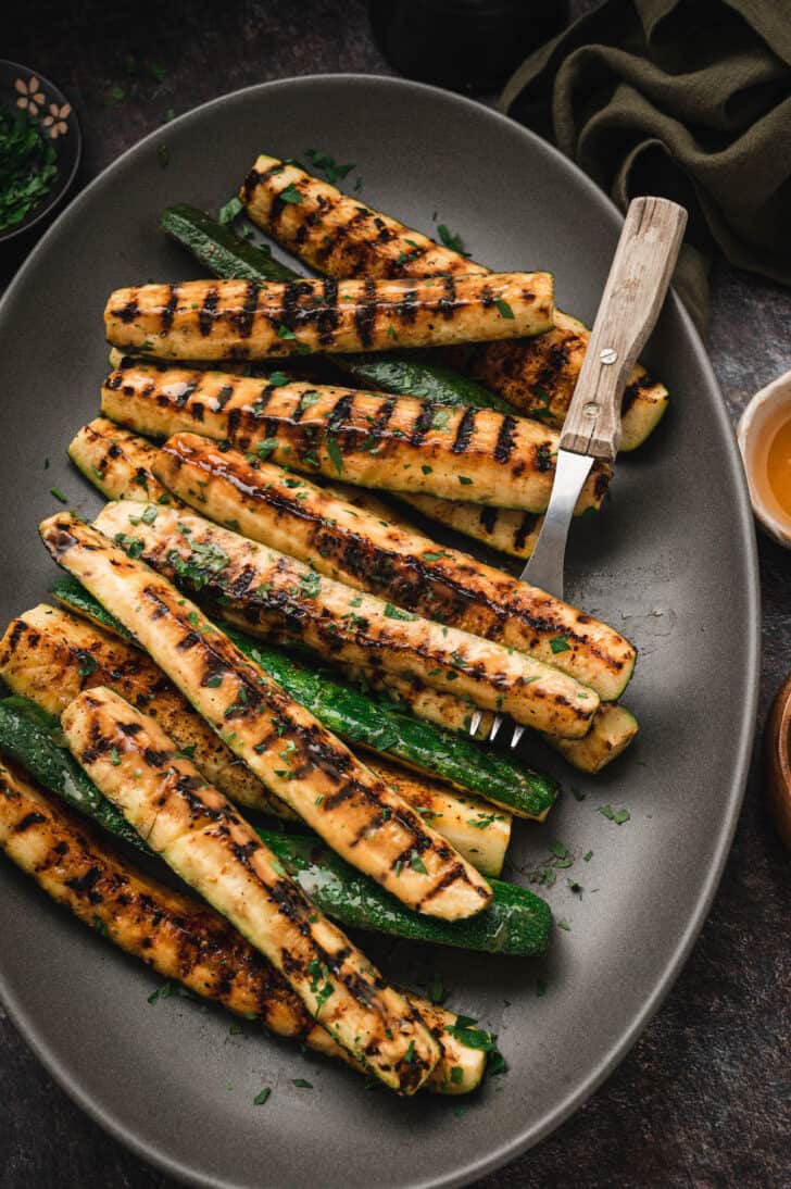 Grilled zucchini spears with a miso glaze on a dark gray oval platter with a serving fork.