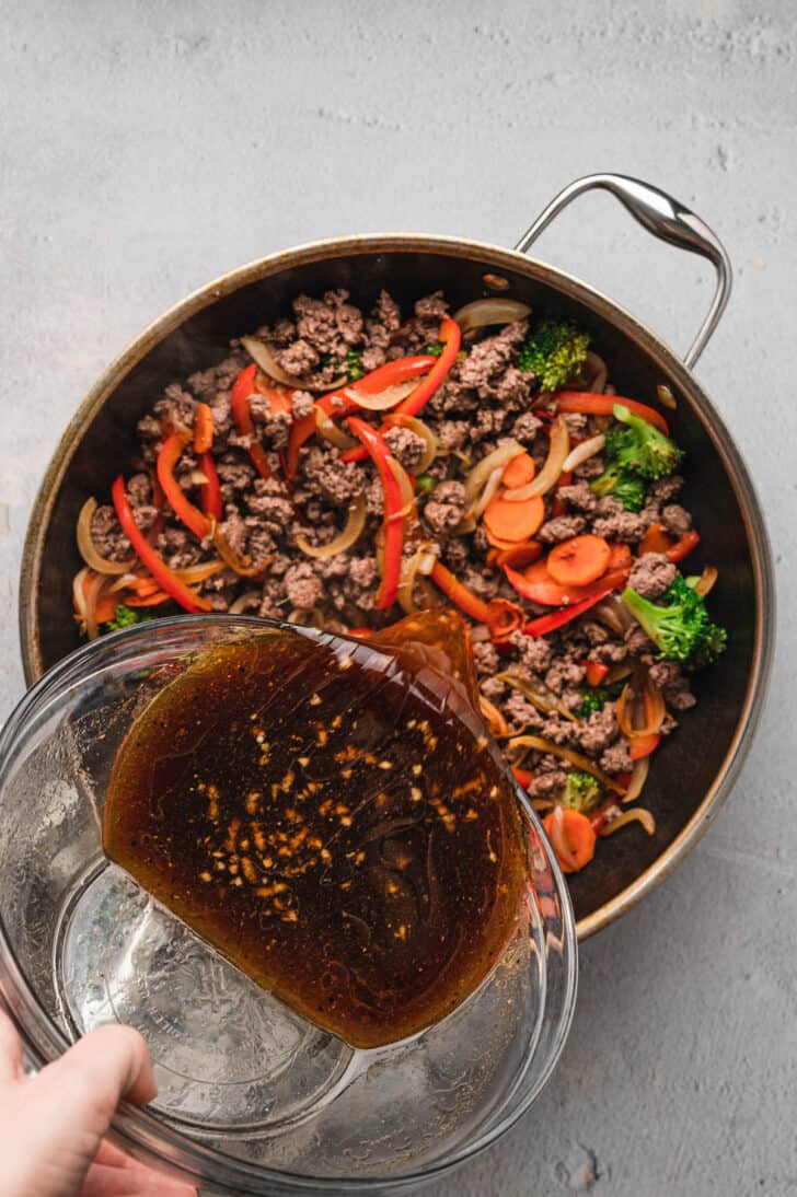 A hand pouring a brown sauce from a glass bowl into a skillet filled with ground meat and vegetables.