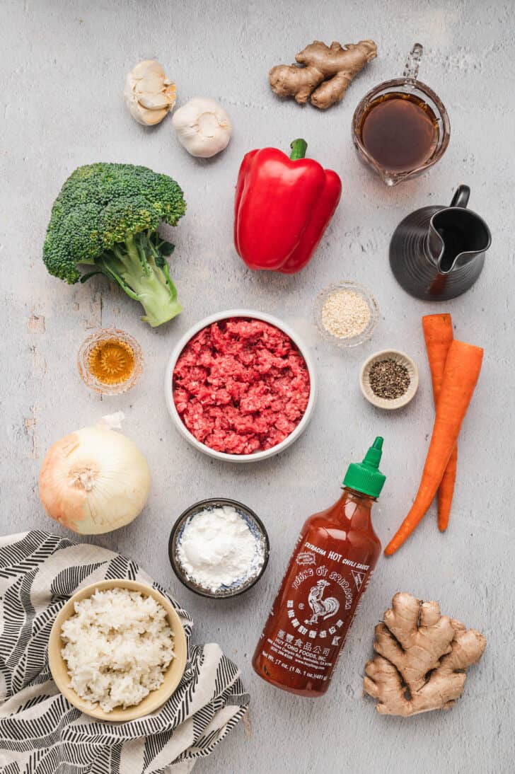 Ingredients laid out on a white surface, including ground red meat, broccoli, red pepper, garlic, ginger, condiments, sesame seeds, spices, carrots, onion, rice and sriracha.