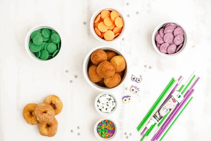 All of the ingredients needed for cake pops for Halloween on a white surface, including green, orange and purple candy melt discs, donut holes and mini donuts, lollipop sticks and candy eyeballs and colored sprinkles.