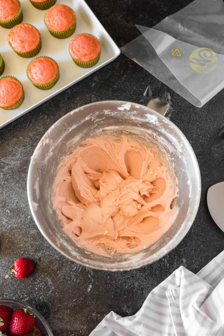 A mixing bowl full of homemade strawberry frosting, with unfrosted cupcakes and a piping bag nearby.