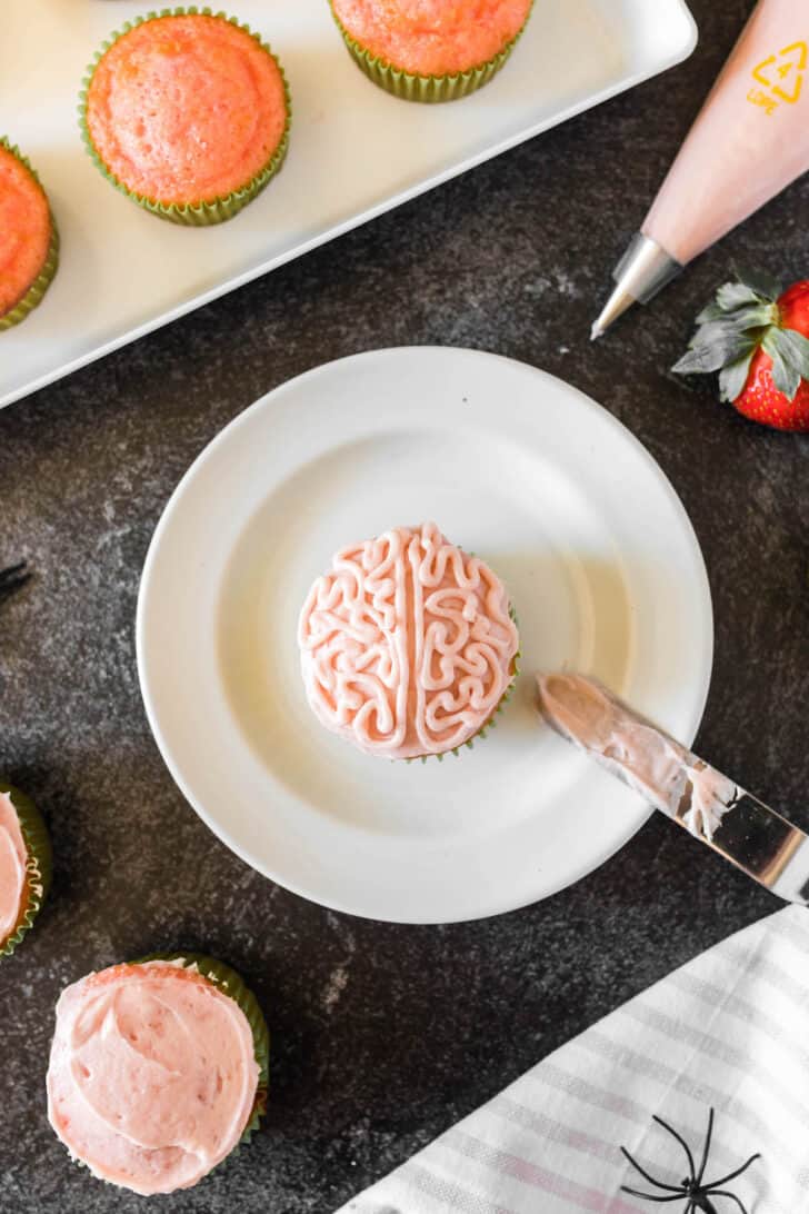 A cupcake on a plate, frosted with light pink frosting, with a zombie brain design piped on top.