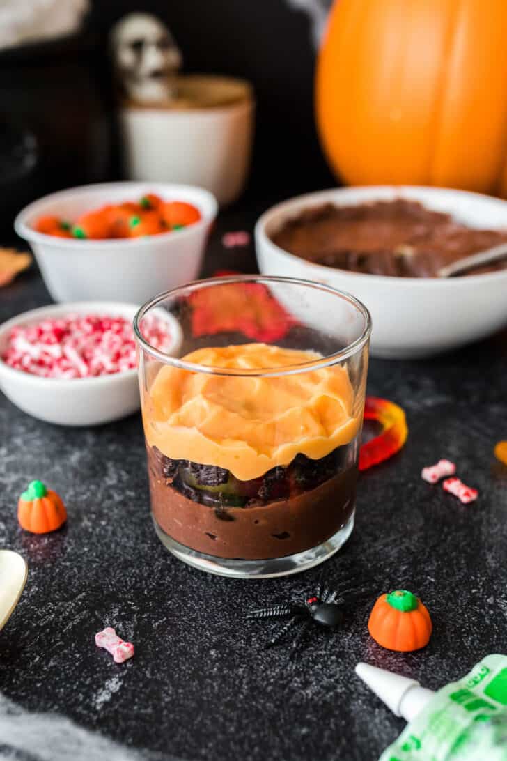 A small glass layered with brown and orange puddings, Oreo cookies and gummy worms.
