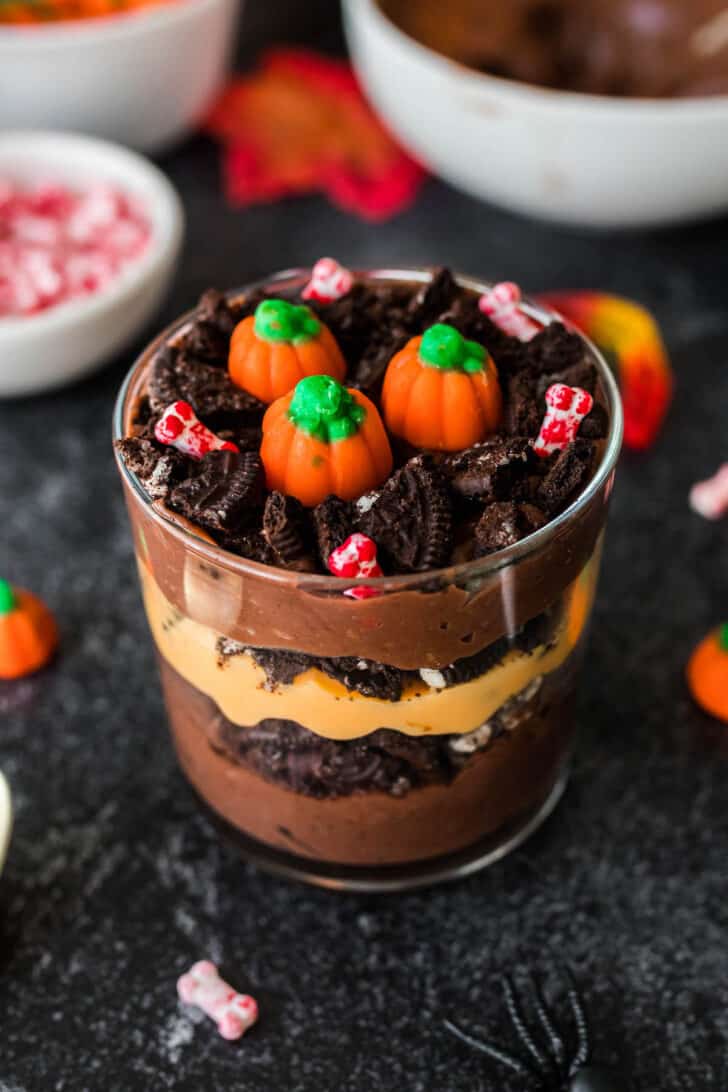 Halloween dirt pudding layered in a small glass. Made with layers of brown and orange puddings, crushed Oreo cookies, gummy worms, pumpkin candies and bone-shaped sprinkles.